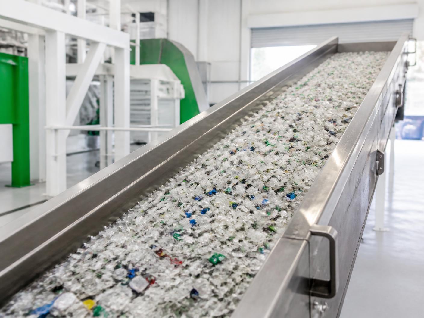 Shredded post-consumer plastic flake on a stainless conveyor inside a bright recycling plant
